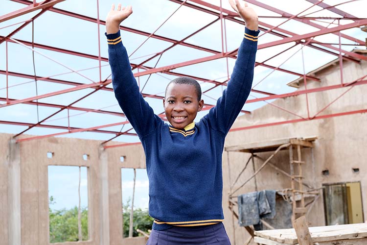 Lucy with her hands in the air with the classroom block construction in the background showing steel trusses.