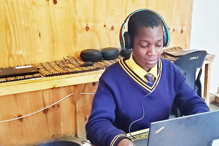 Student Evelyn is seating typing on a laptop with headphones on listening to an English lesson. 