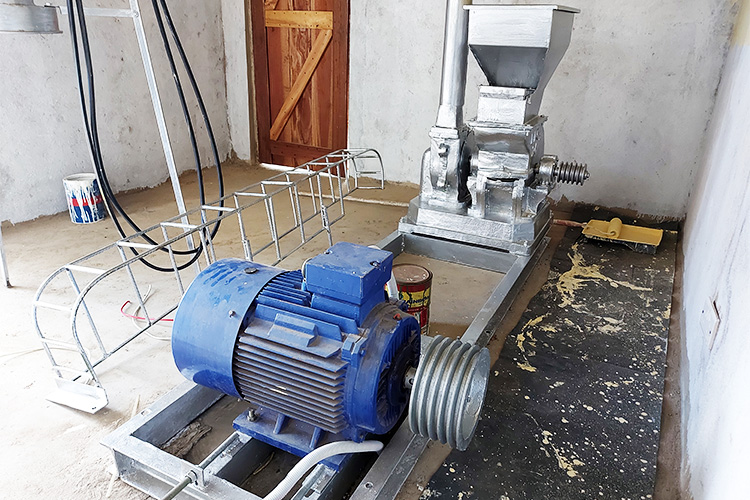 Inside of the maize grinder mechanical room showing the electric motor, conveyor belt assembly and the hopper in the background.