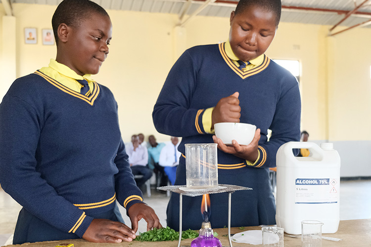 Two Kuwala students with a bunsen burner in the foreground and the second student working a mortar and pestle.