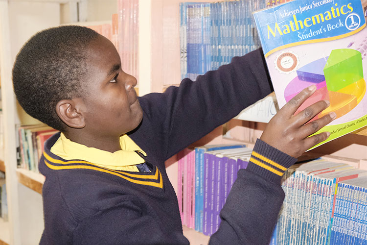 Sharon, a Kuwala student is in the library putting back a math workbook on the shelf.