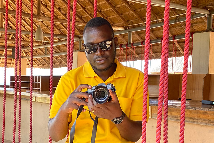 Chembekezo inside open frame building holding Fujifilm camera with ropes hanging from the ceiling.